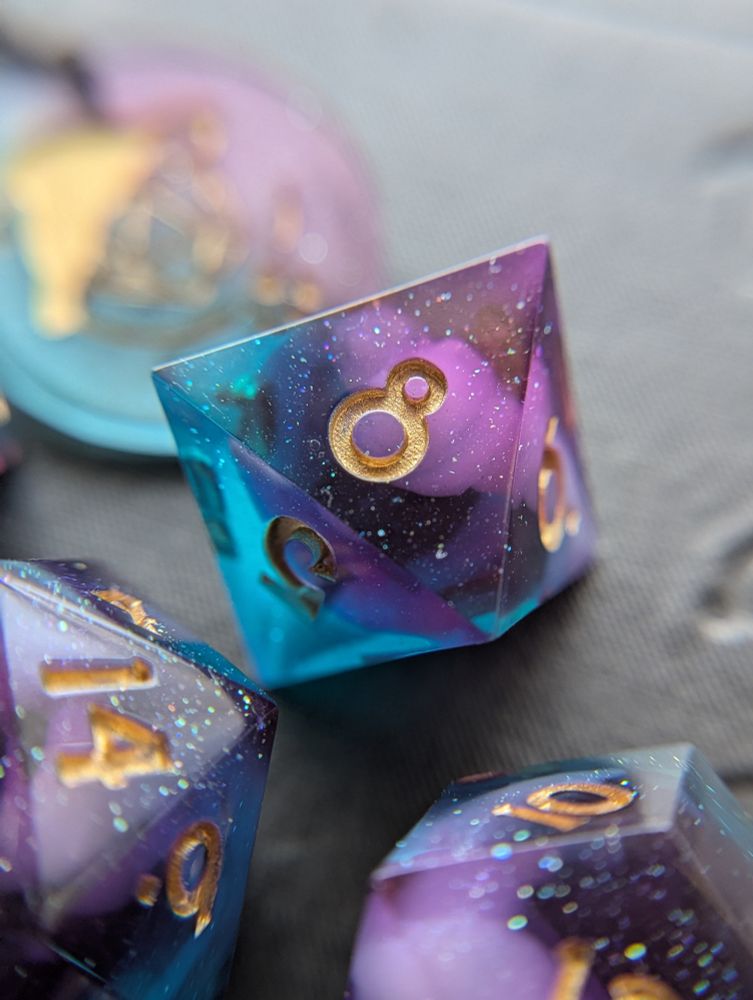 Handmade dice with white swirls in purple and teal transparent resin. They are inked gold.