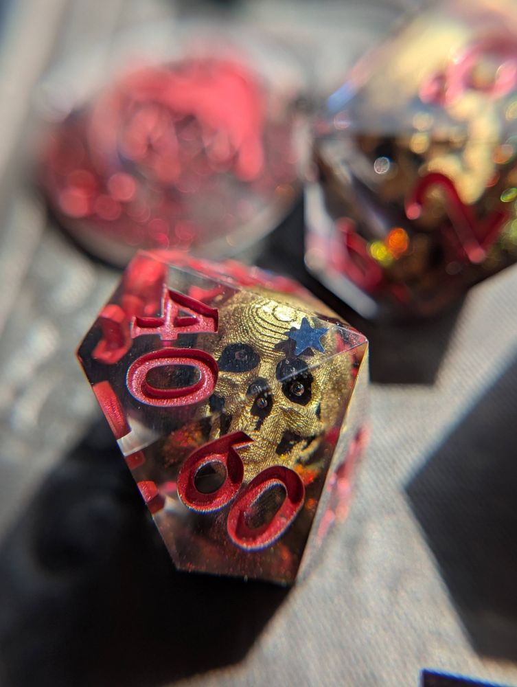 Handmade dice featuring gold 3D printed skulls and swirls of red/green/yellow chameleon pigment, as well as black holographic stars.