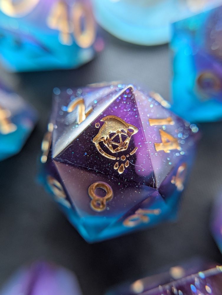 Handmade dice with white swirls in purple and teal transparent resin. They are inked gold.