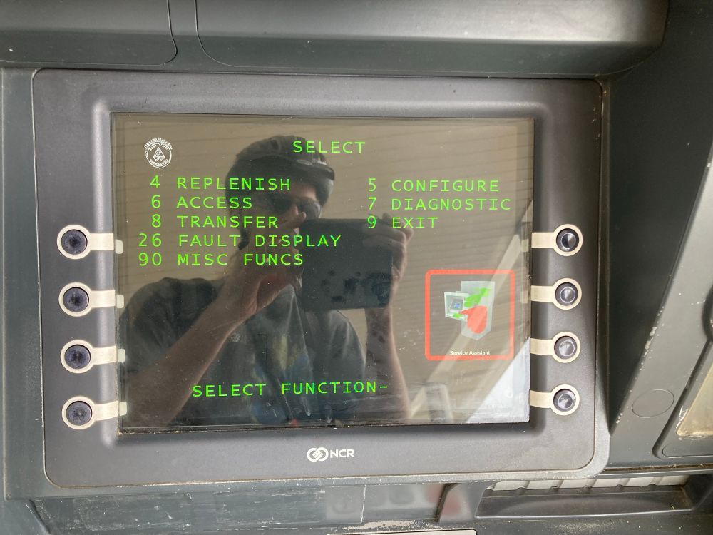 ATM screen showing a menu with options: REPLENISH, ACCESS, TRANSFER, FAULT DISPLAY, MISC FUNCS, CONFIGURE, DIAGNOSTIC, EXIT. The reflection shows me wearing a bike helmet and sunglasses taking the photo