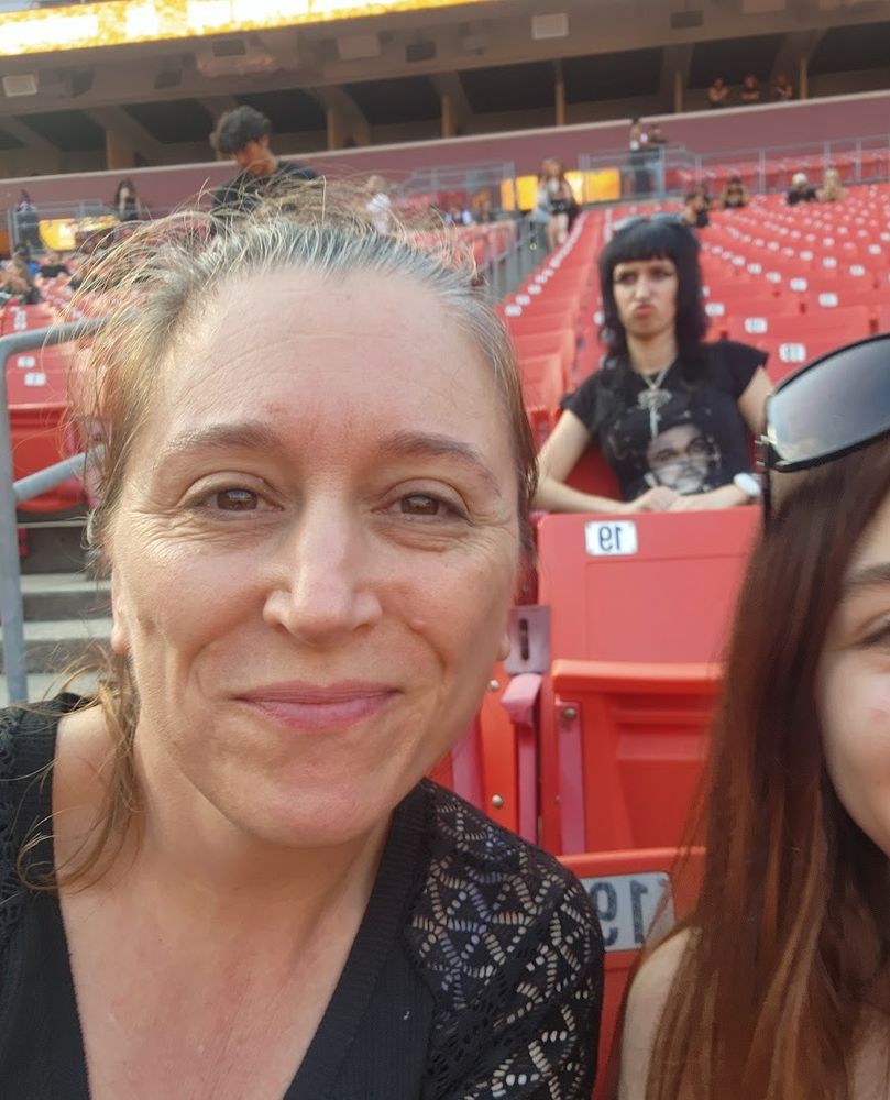 Me sitting in a mostly empty set of seats in a stadium. My brown hair is back in a ponytail and I'm wearing a black shirt with lacey shoulders. Behind me is a goth girl with black hair and a black t-shirt who I accidentally caught mid-sneer/smooth. She's a goddess.