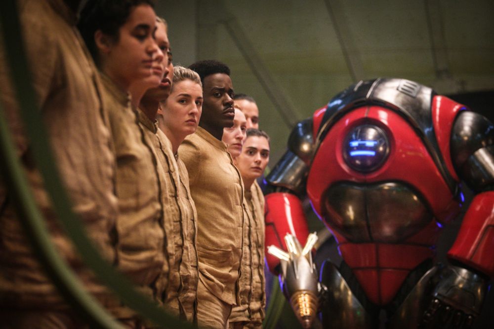 A scene of Doctor Who's Robot Revolution depicting the doctor standing lined up with other people, all wearing beige outfits. A grumpy looking giant red robot looms in the background, carrying a laser gun in his right hand. The doctor looks serious towards the camera. 