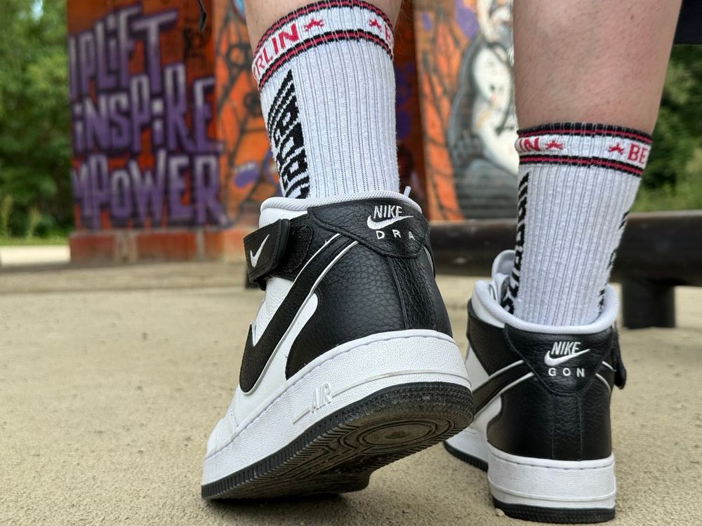 A photograph of the same sneakers on feet from behind. The heel is black, the left heel having letters spelling "DRA" and the right having "GON" on it. The graffitied wall can still be seen in the background, spelling something like "lift", "inspire" and "power".