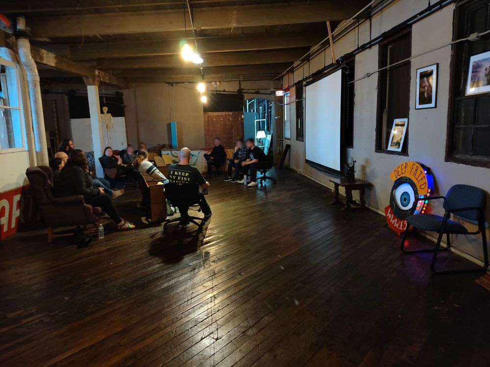 A group of people sitting in a circle after the screening, discussing the film. The room is an old warehouse with wood flooring, yellow lights, and a projector screen.