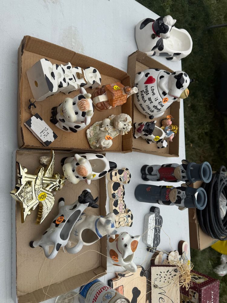 A white folding table has numerous cardboard boxes being used as display trays. you can see multiple black and white cow figurines of various shapes and sizes 