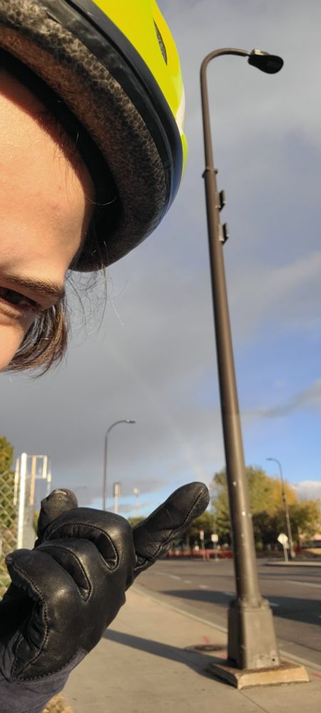 A faint rainbow seen from the street, and Rebekkah's hand positioned just so that it looks like the rainbow is coming out of her finger. Rebekkah's face is peering out from the left side