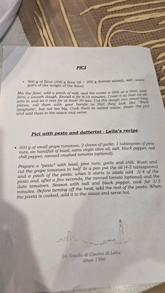 A printed recipe page titled “Pici,” placed on a kitchen counter beside long rolled strands of dough. The text lists ingredients and instructions for making pici pasta and a tomato–basil pesto sauce. At the bottom of the page is a simple line drawing of a town skyline with the words “La Scuola di Cucina di Lella since 1996.”