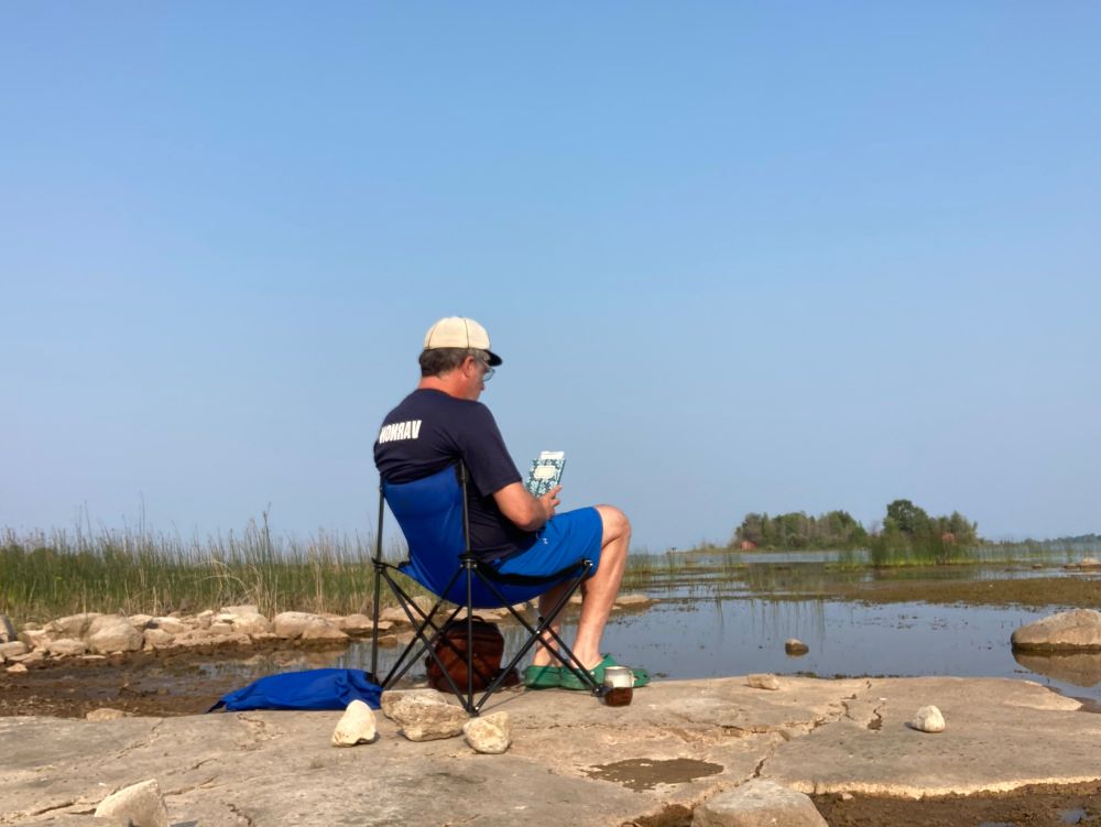 Man in ball cap sitting in chair on rocky beach at Little Red Bay, with Sunset Island in the distance.