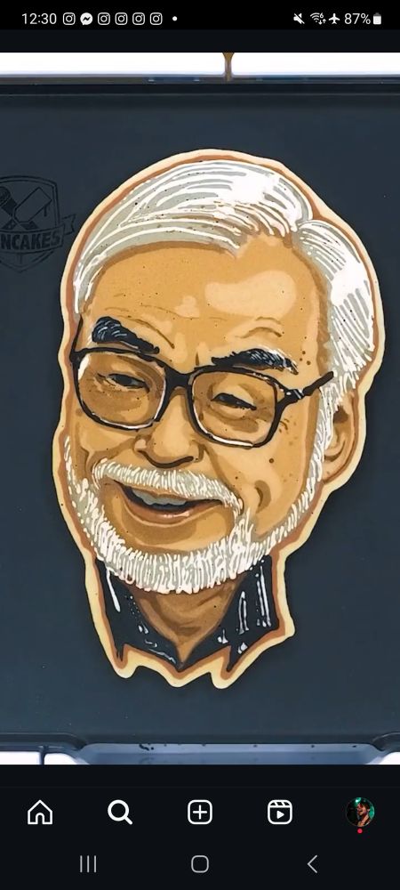 Pancake art portrait of Hayao Miyazaki, featuring intricate shading and color detail. He is depicted with white hair, a matching beard, black-rimmed glasses, and a warm smile. The pancake is shaped to match the contours of his head and shoulders, displayed on a black griddle.
