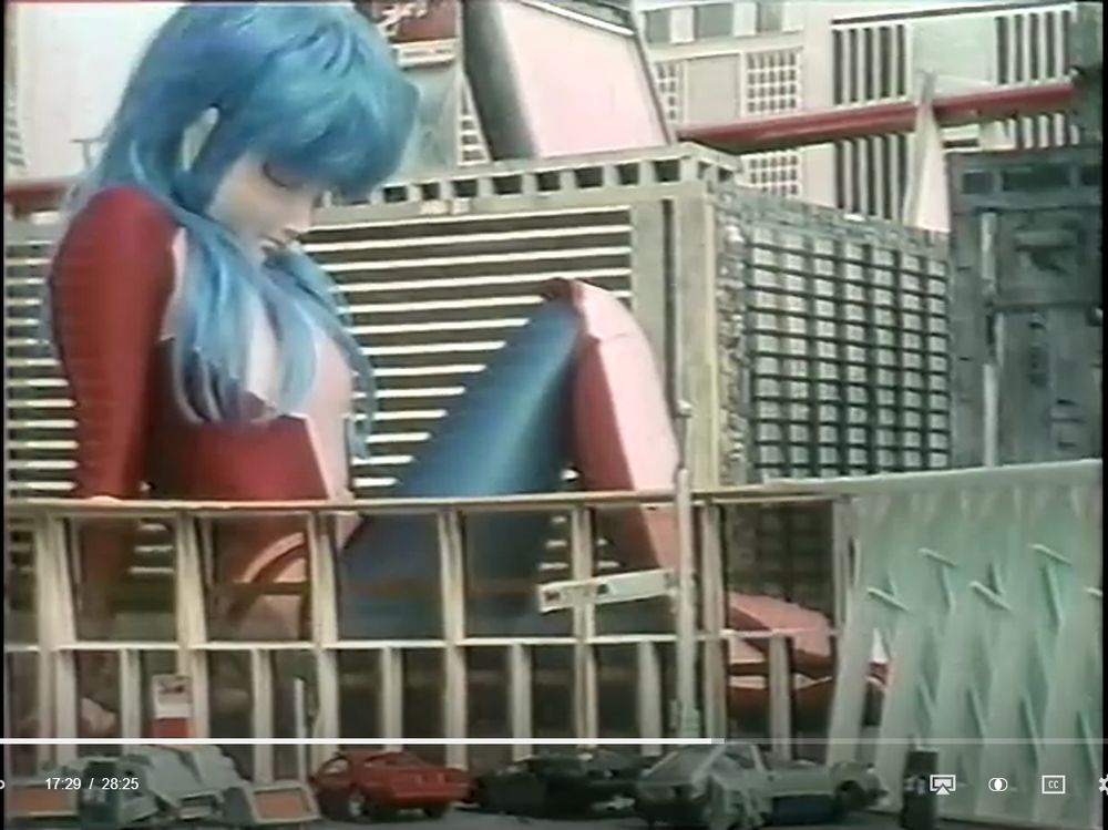 Still-image from the 1984 Japanese independent tokusatsu film "All About Mighty Lady". A large, blue-haired giant woman in a mask and bodysuit sits amid city buildings, surrounded by cars and infrastructure.