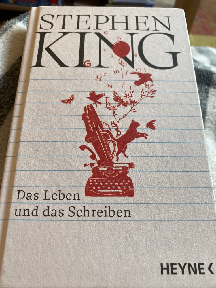 Cover von Stephen Kings „Das Leben und das Schreiben“. Cremeweißer Hinter Grund mit feinen blauen Linien, die an ein Schreibheft erinnern. In schwarzer Schrift oben zentriert  der Autorenname, ziemlich weit unten linksbündig der Buchtitel, ganz unten rechts der Verlag. Mittig in roter Farbe stilisiert eine Schreibmaschine, aus der ein Auto, eine Katze sowie ein Zweig, Schmetterlinge, Vögel, ein Luftballon und Buchstaben springen. 