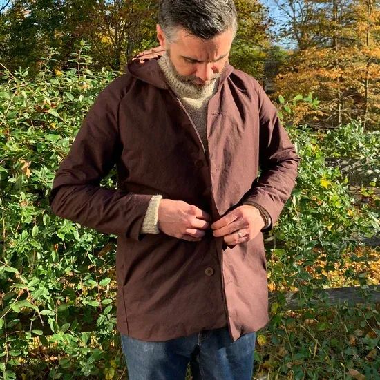 A man looks down as he buttons his brown coat. It has a darted saddle sleeve, a generous hood, deep pockets, and a relaxed fit.