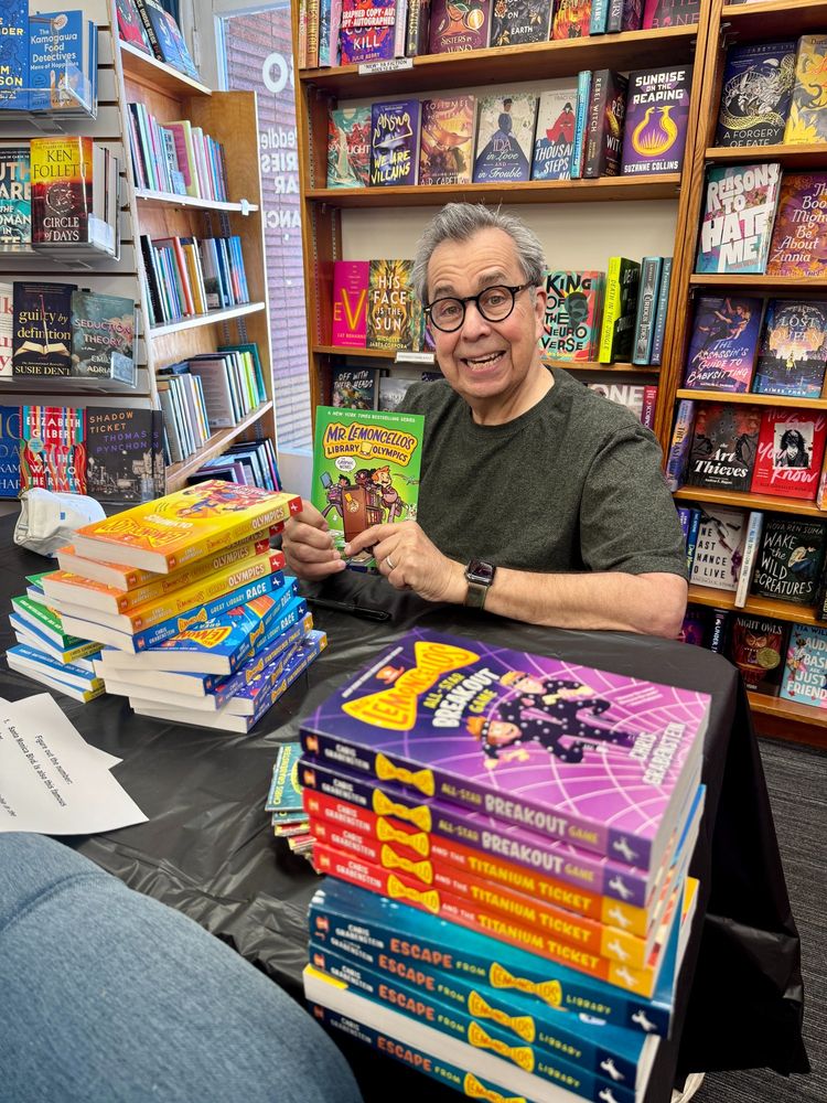 Chris Grabenstein holds up a book and sits among many stacks of his books.