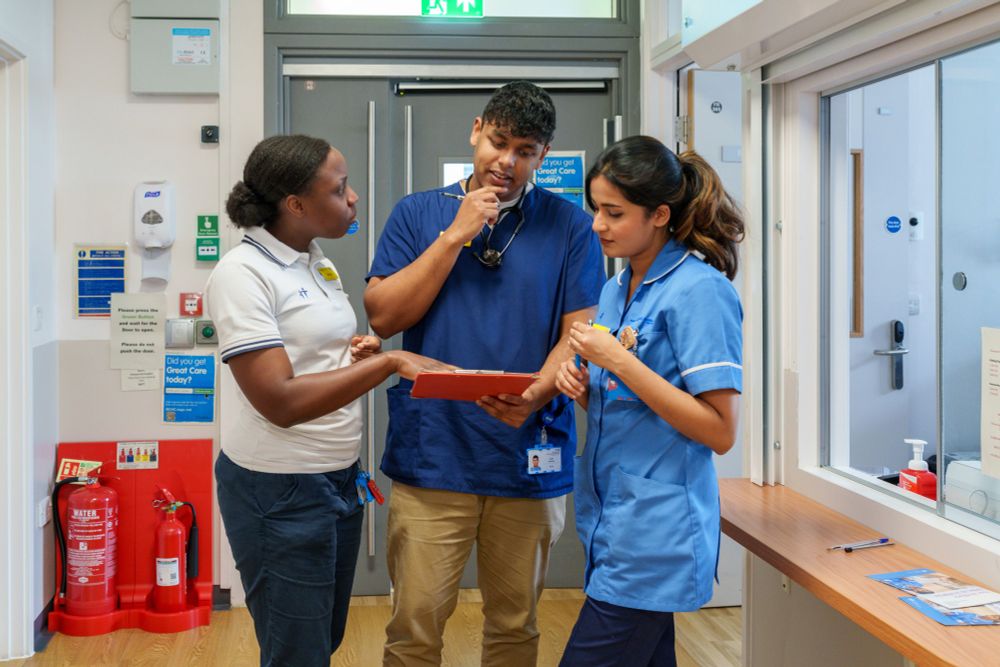 Three health care workers standing together in a hospital corridor and discussing a paper document.