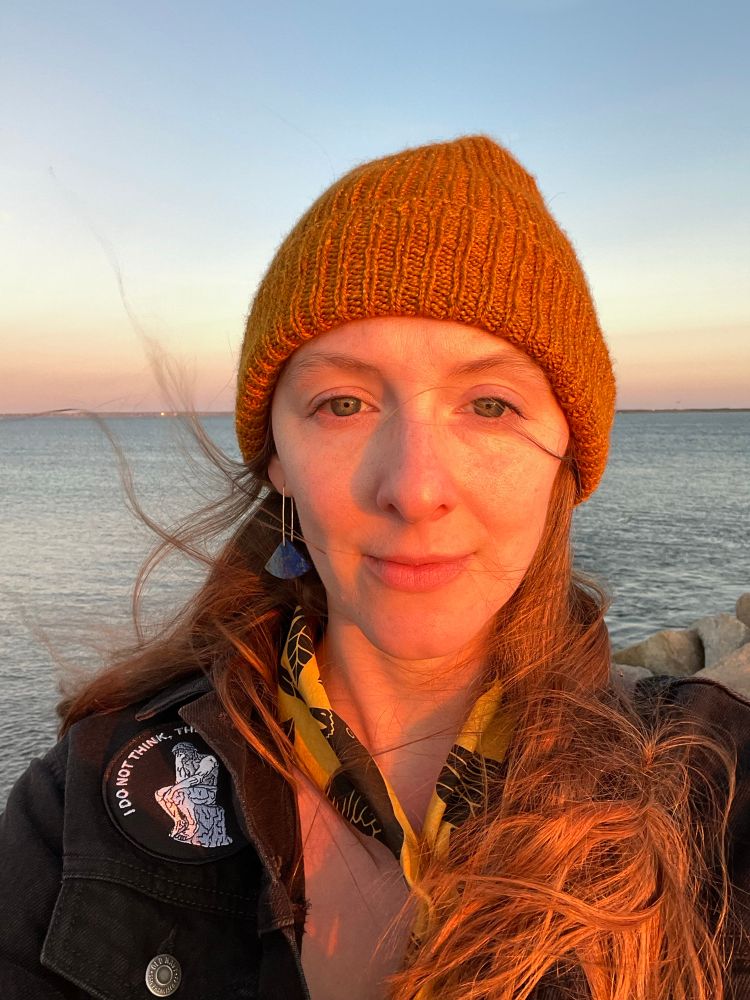 Portrait of the artist, a white female with red hair wearing a beanie, at sunset with water behind her. 