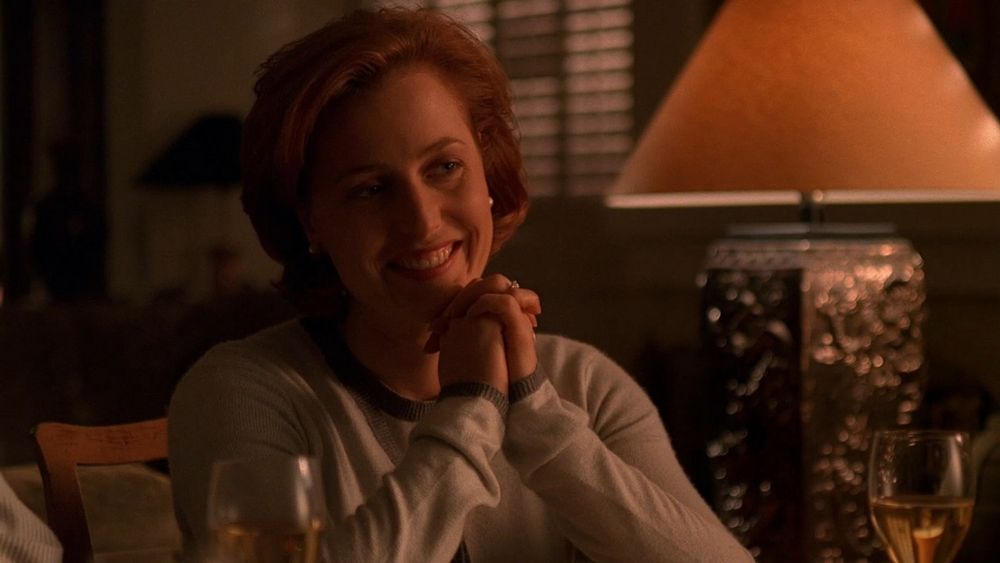 Screenshot of Scully from The X-Files dresses conservatively, sitting at a table and smiling. 