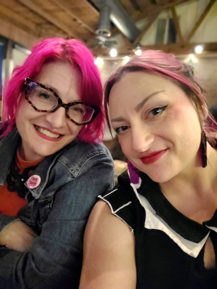 Two femme presenting people with different shades of pink hair sitting next to each other and smiling. 