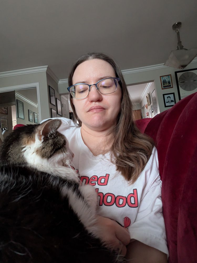 A white person with shoulder length brown hair has the cutest cat in the world curled up on their belly and chest
