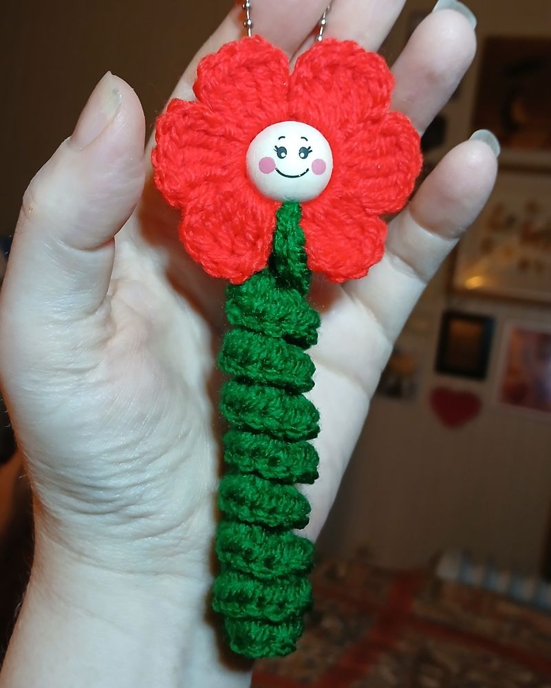 A woman's hand holding a crocheted red flower with a wooden bead face and a green spiral stem