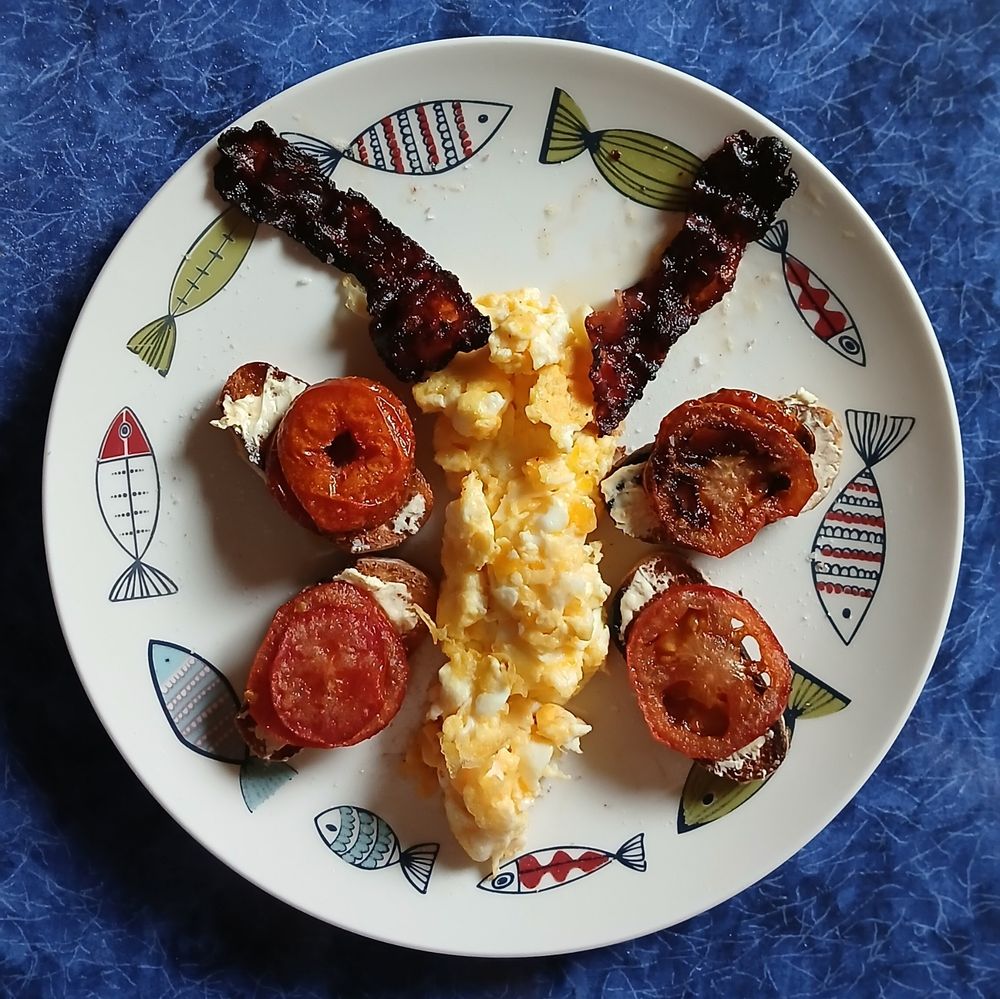 A plate of scrambled eggs , bacon , toast and tomato slices arranged in the shape of a butterfly
