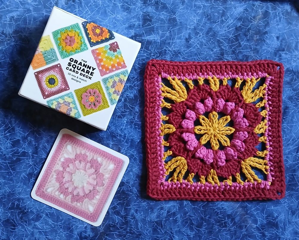A box labeled "GRANNY SQUARE CARD DECK," a card from the deck showing a black and green coloured granny square, and a crocheted granny square as depicted on the card in red, pink, and gold