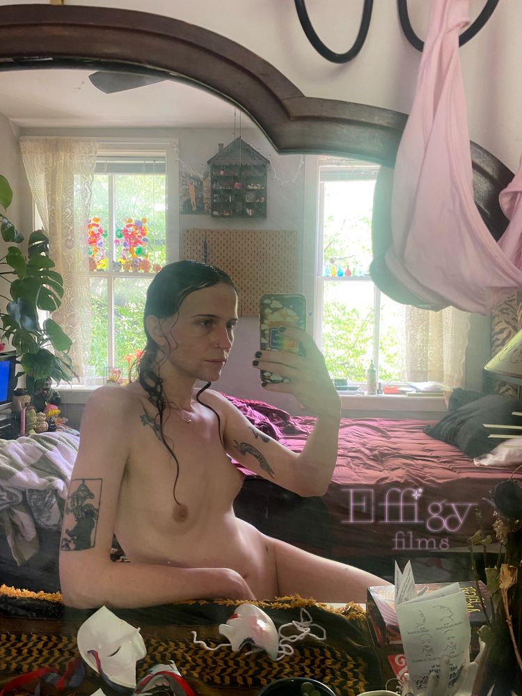 Aura sits at a desk and takes a picture in her new mirror. She leans on one arm that rests against the desk which is covered in a yellow and black keffiyeh. A broken clown mask sits on the desk along with various items, a zine in a porcelain hand, 2 stacked VHS's. Her other arm holds up the phone, which she is looking at. Her hair is wet and her body is in half-profile, the light from the windows behind her highlight the curves of Aura's body from shoulder down to her leg which stretches out below the desk. The bed behind her is unmade, it sits across from a monstera plant and a tv with a blue screen. 