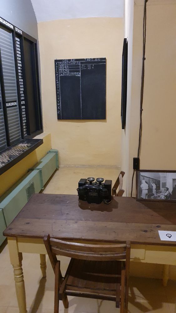 Ein enges Zimmer. Im Vordergrund ein einfacher Tisch, auf dem nur ein schwarzes Telefon steht und ein Foto von dem Raum, als noch benutzt wurde. Hinten eine Tafel. Links eine Einstecktafel, in dem der Zustand verschiedener Squadrone gekennzeichnet werden kann. Details: Foto: ein Mann sitzt am Tisch mit übereinander geschlagenen Beinen und hat den Hörer am Ohr. Zwei Männer stehen an der Einstecktafel. Tafel: In der linken Spalte A, D, E, G, H, J, K, M, O, P, Q, R, S, U, V, W, X, Z. In der mittleren, OPS AIC, in den ersten fünf Zeilen Foodstuff 30, BR24, BR30, BR38 und 4BONAJMFF(30), in der rechten Spalte TEST AIC in der Zeile D SL 22 7, Zeile E SL 10 4.