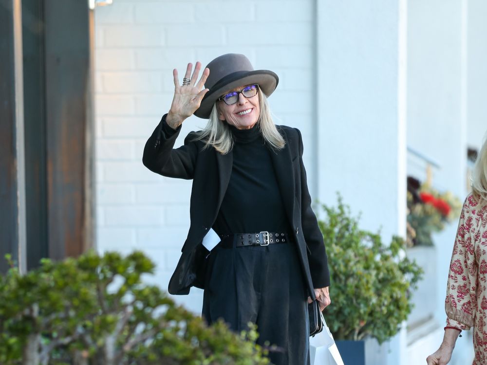 Diane Keaton, stylishly dressed in grays and blacks and wearing one of her large brimmed hats, waving and smiling outside on a sidewalk.