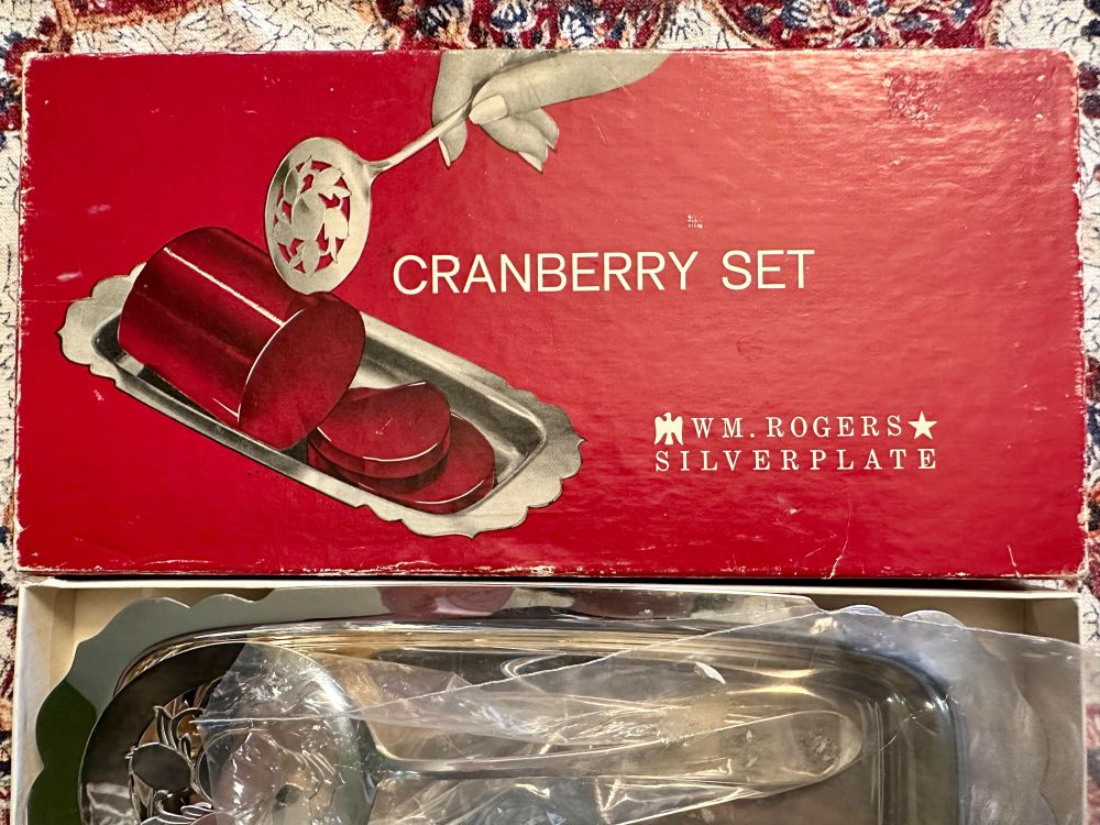 The box top of the W M Rogers silverplated canned cranberry sauce server tray and slicer utensil. The box displays a female hand holding the utensil and slicing through cranberry sauce on the silver tray.