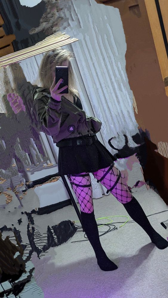 A mirror selfie of Tmiah standing wearing an olive green jacket, half zipped and one shoulder off, revealing the black crop top and leather chest straps underneath. Further down is her black mini-skirt, with leather thigh straps and fishnet leading to her black thigh socks.