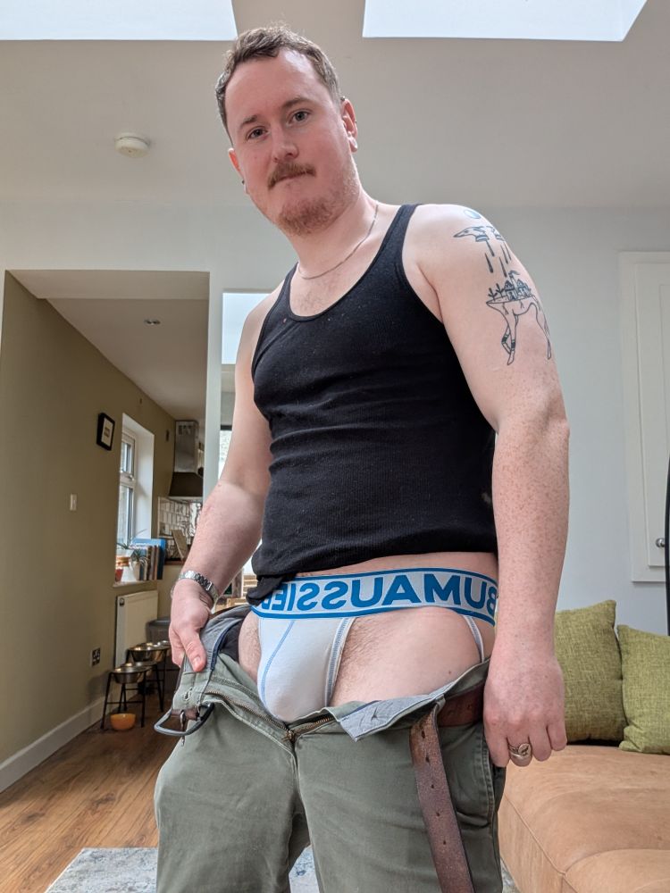 White man in room with natural light wearing green trousers, white jockstrap, black vest, looking down towards camera