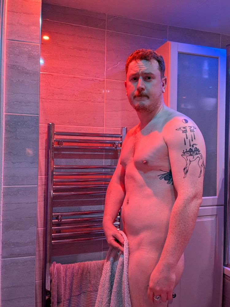 White man in bathroom holding towel in front, lighting is orange and blue 