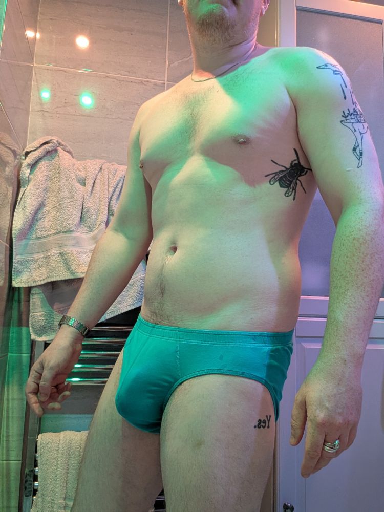 White man in bathroom, green and orange lights, wearing blue underwear 