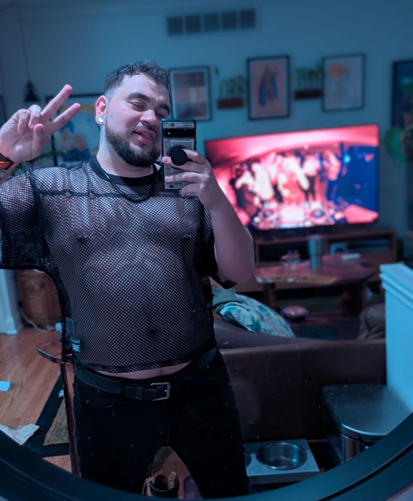 Selfie of me in black pants and a black mesh shirt. You can see my nipple rings. Living room in the background, tv playing disco. 