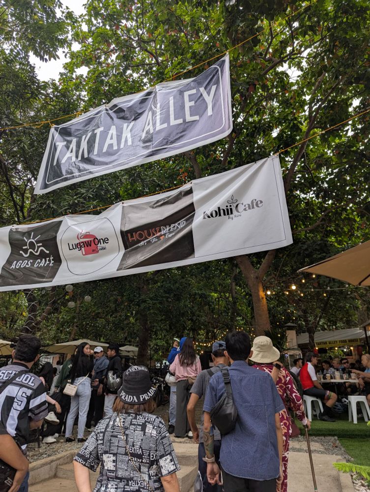 A crowd walking into Taktak Alley under tree shade, with banners showcasing various café logos including "Agos Café," "Lugaw One," "House Blend," and "Kohii Café."