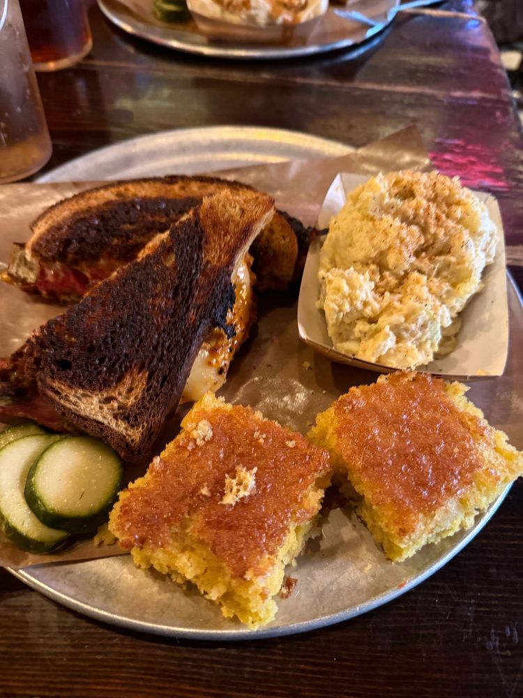 A plate of food. It includes a smoked corned beef Reuben sandwich, potato salad, two pieces of corn bread, and a few slices of pickles.