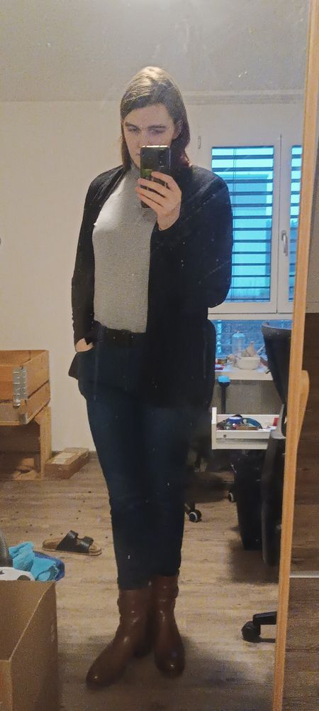 picture of a white woman with brown hair, dressed in brown ankle boots, dark blue slim jeans, a grey turtleneck, a black overshirt, taken in a mirror