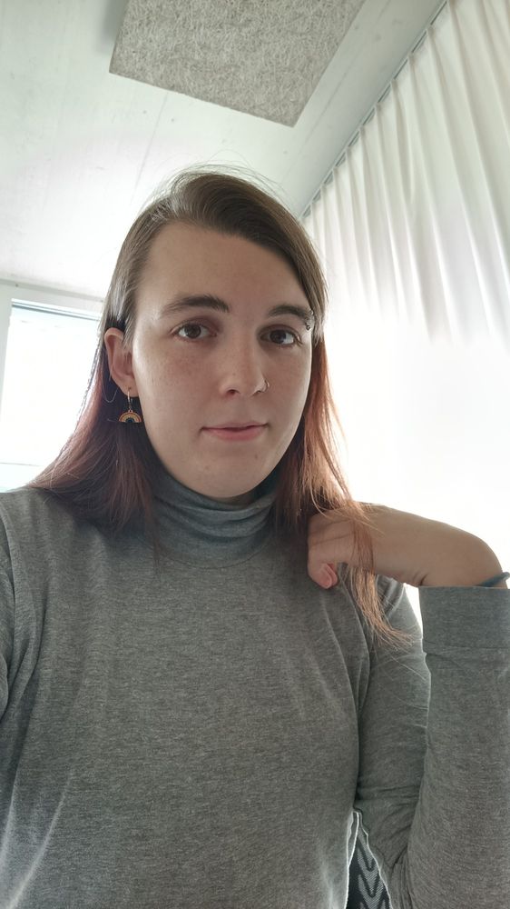 me in a grey turtleneck and rainbow earrings against a sunny window background, left hand half curled up against my shoulder, with a few strands of hair falling in front of it