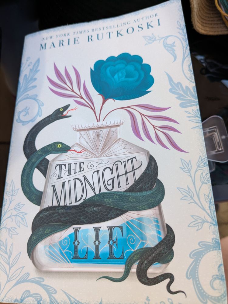 The front cover of Marie Rutkoski's The Midnight Lie. The picture depicts a bottle containing a blue flower with pink petals. Curled around the bottle are two snakes. Flower motifs surround the edge of the cover. 