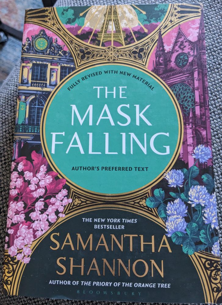 Front cover of Samantha Shannon's The Mask Falling, book four in her Bone Season series. The colour scheme of the cover is predominantly pink and turquoise with gold accents. Contains a stylised depiction of the Eiffel Tower and the signature flowers on the cover. 