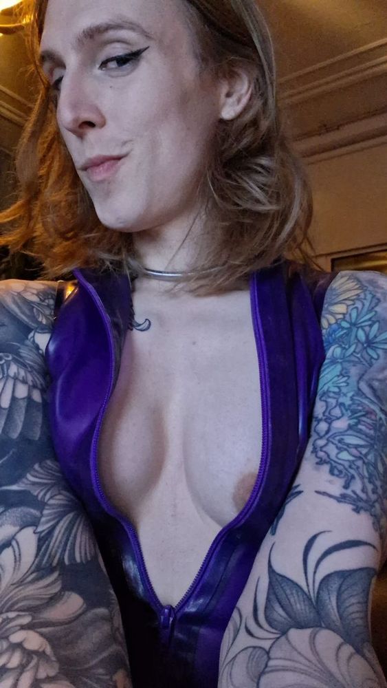 Calliope, in a mauve latex front zip minidress, zipper pulled down, exposing its tits. A touch of nipple showing. It wears a silver collar