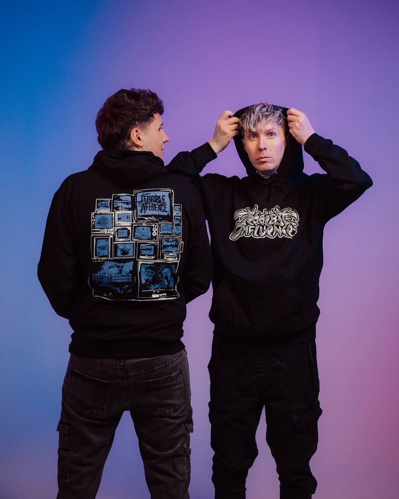 Dan and Phil standing side by side. Dan has his back to the camera showing off the back design of the tour hoodie. he is looking across to the side at Phil who is facing the camera and is holding the hood of the hoodie up with his hands