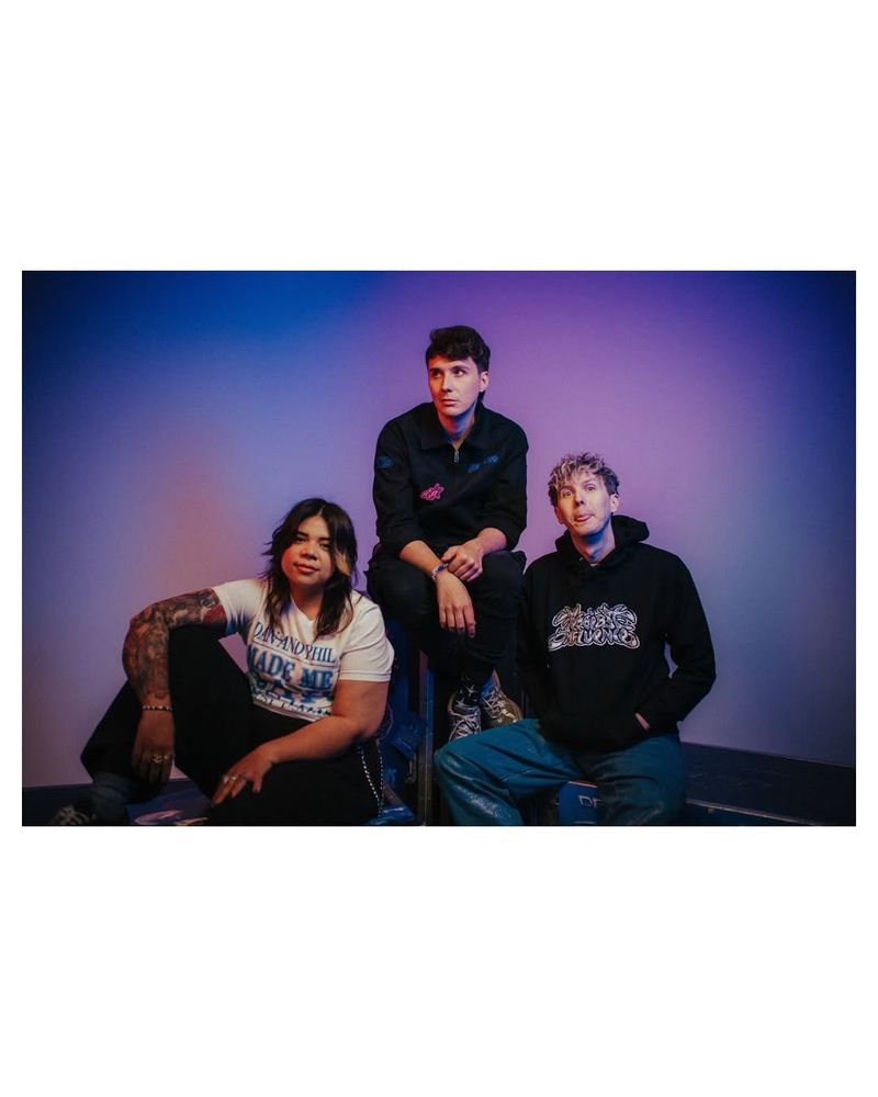 a photo of Dan, Phil and Sarah (merch team). Sarah is on the left looking at the camera wearing the "Dan and Phil made me gay t shirt". Dan is in the middle wearing the tour jacket with his hands crossed and looking to the side. Phil is sat on the right wearing the tour hoodie, his hands in his pockets and sticking out his tongue