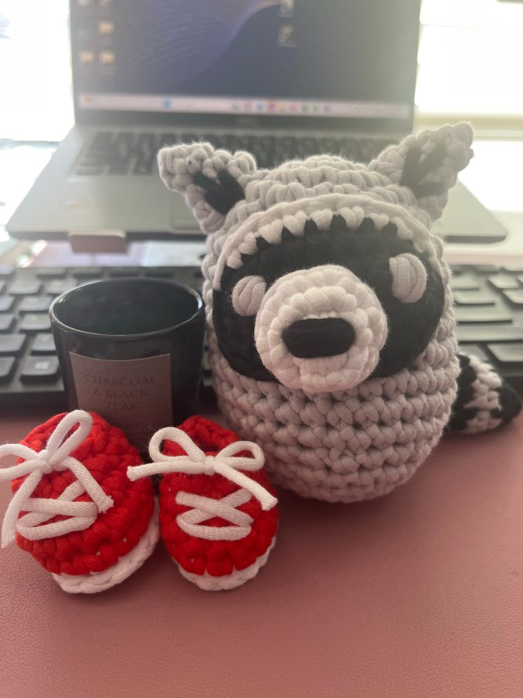 a crocheted raccoon with slightly askew features, propped up against a small candle. little red crochet shoes with tied laces placed in front of the candle.