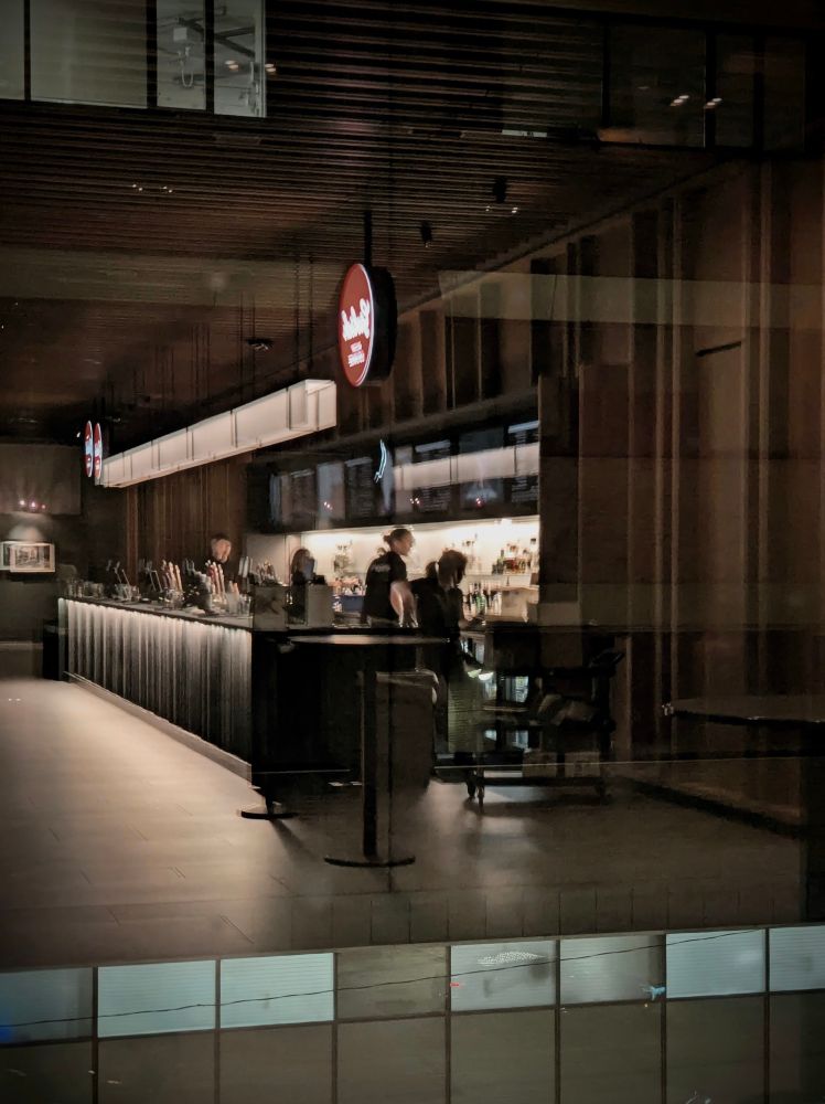 A photograph of the west facing wall and windows of St Michael's Hospital shot from the balcony level bar of Massey Hall. The long bar counter is free of patrons during a performance.  Four staff members are busy with other tasks. A long rectangular light hangs  between two red circular signs above the counter.  The longest wood panels and details of the walls and ceiling blend into the stark rectangular frames of the hospital windows.