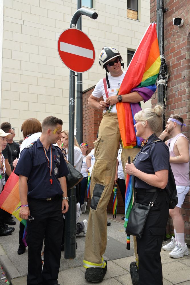 A photograph of a group of three fire and emergency workers in front of a crowd gathered for the Pride Parade. The man and woman at the front are in a regular uniform of dark trousers and a short sleeved button up. The third member of the group is on stilts making him as tall as the no entry sign behind him.  He's wearing khaki fire fighter pants with red suspenders, a white t-shirt, black and white helmet, and dark glasses.  He is also holding a pride flag.