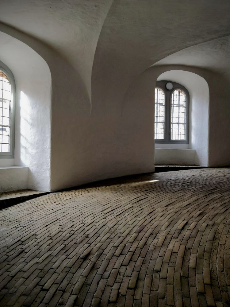 A photograph of the spiraling interior ramp of Copenhagen's Rundetaarn (Round Tower).  The pathway is constructed from interlocking brick in portrait orientation. (There's probably a better bricklaying word for that, but, alas, I am not a bricklayer.)  The windows are pairs of long, narrow arches inside arched alcoves.  An arch also runs inward from the wall between the windows to the centre of the tower.  The walls have been parged with a warm white stucco (again, possibly the incorrect term).  The space is old (17th century), but somehow still manages to have the minimalist structure and colouration one associates with modern Danish design.