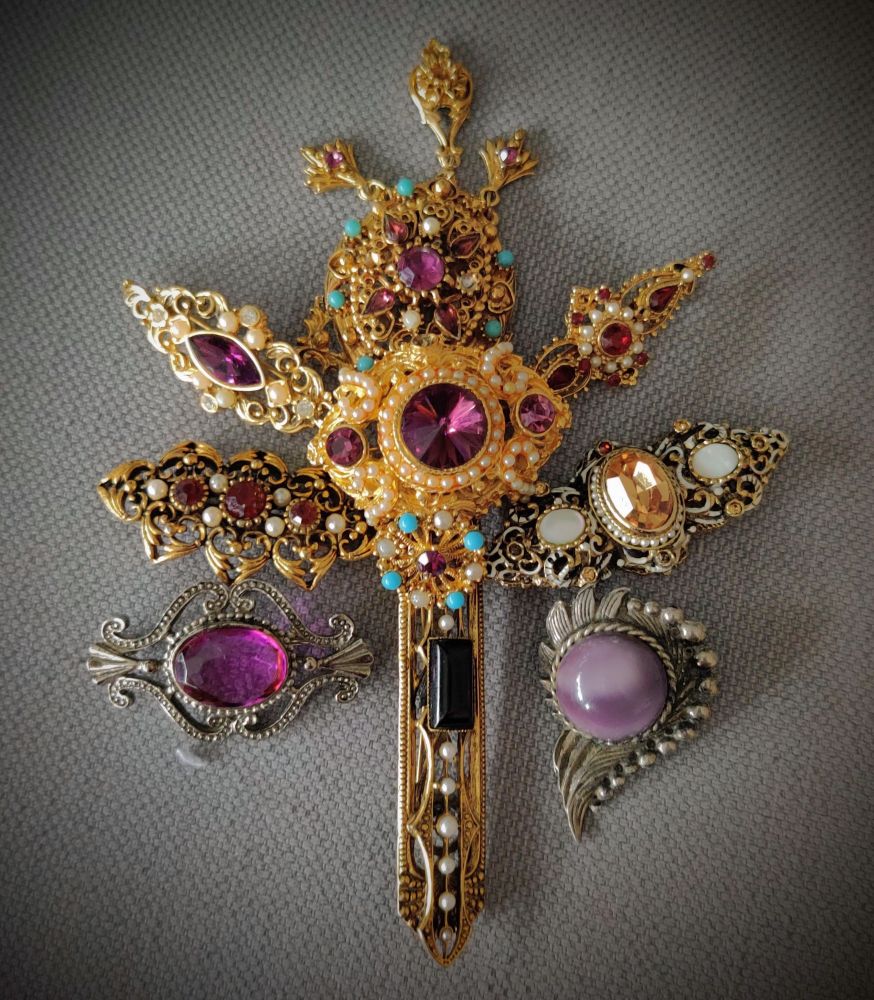 An assemblage of ten vintage brooches on a grey background. 

The brooches are in a mask-like configuration resembling two eyes, a nose piece, and a head dress.

Brooches in the head dress and nose piece are gold tone metal.  The two used for eyes are silver tone.  Gems are predominantly purple with small turquoise beads and white pearls.  There is also one large black stone on the nose piece and a large amber crystal in one of the head dress fans.