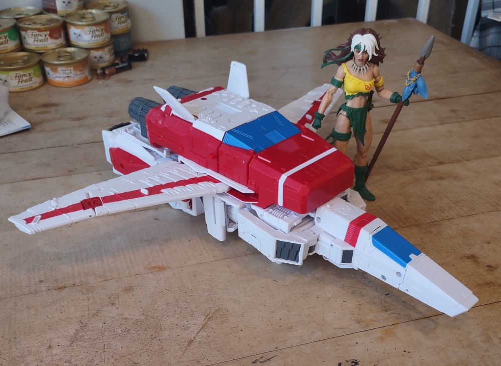 A.6" Rogue action figure stands on a wood table next to a large white and red toy jet plane. The jet is the size of a car compared to her.