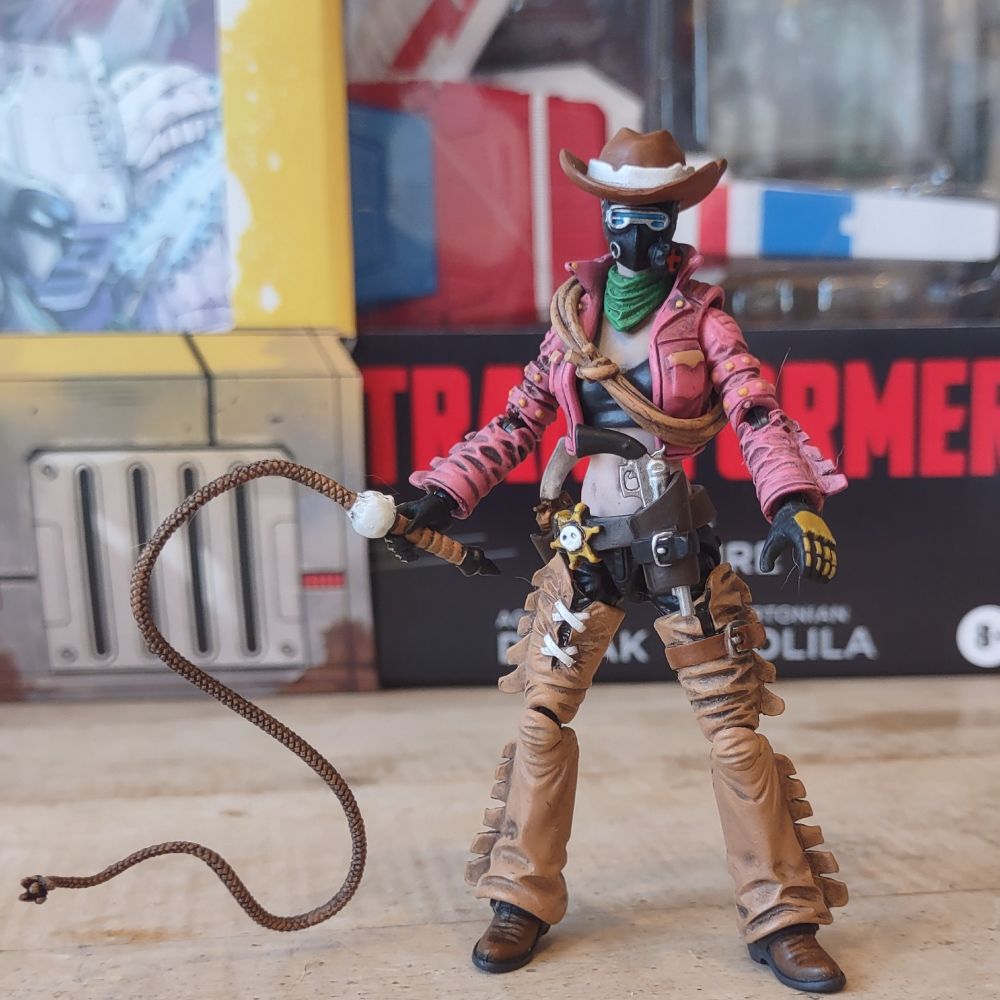 An action figure of a  gas-mask wearing woman in a cowboy hat and pink leather jacket. Her whole aesthetic is sort of post-apocalyptic cowgirl. She's got a long leather bullwhip in one hand.